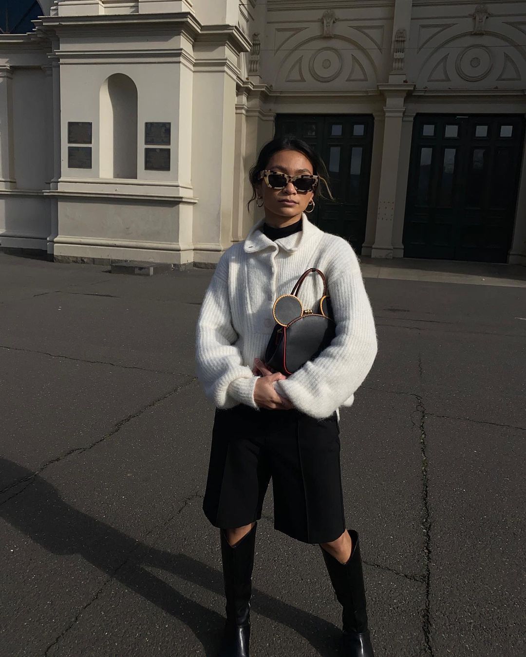 Woman in white sweater and black shorts and boots stands in front of building. She has sunglasses and is holding a black purse.