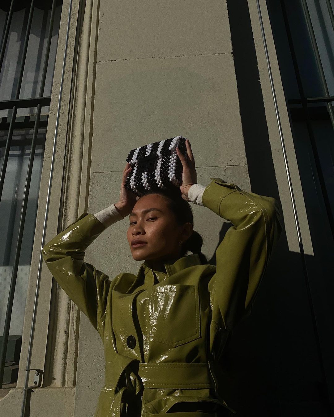 Adult female in a glossy green trench coat poses with a beaded black and white bag above her head in front of a light grey building.