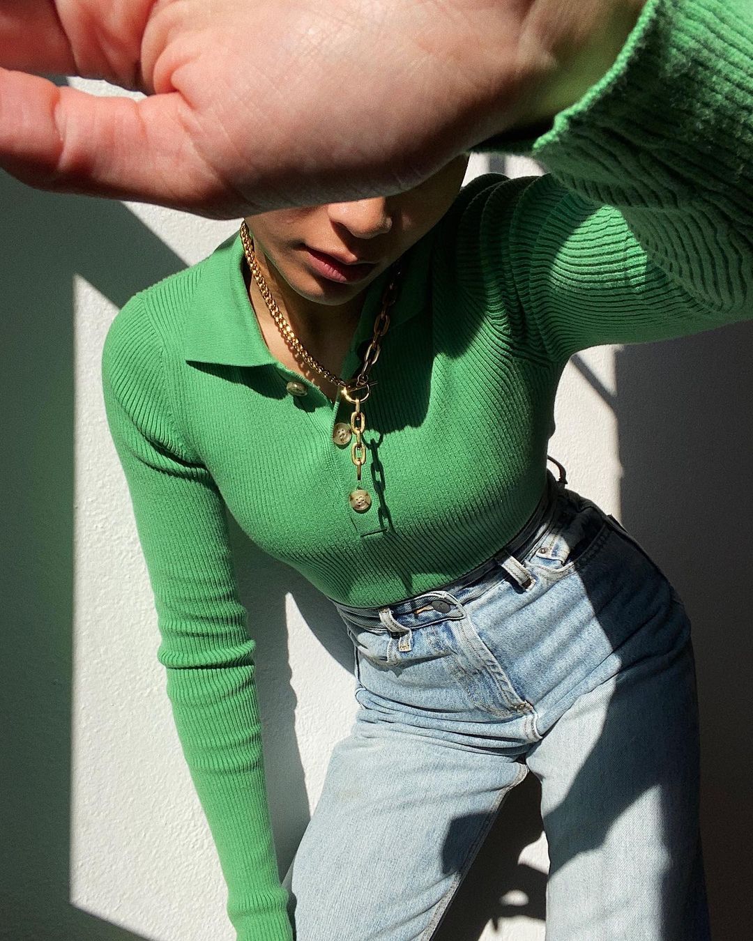 Woman in bright green top and jeans shielding face with hand. Gold necklace and belt add detail to the fashion portrait.