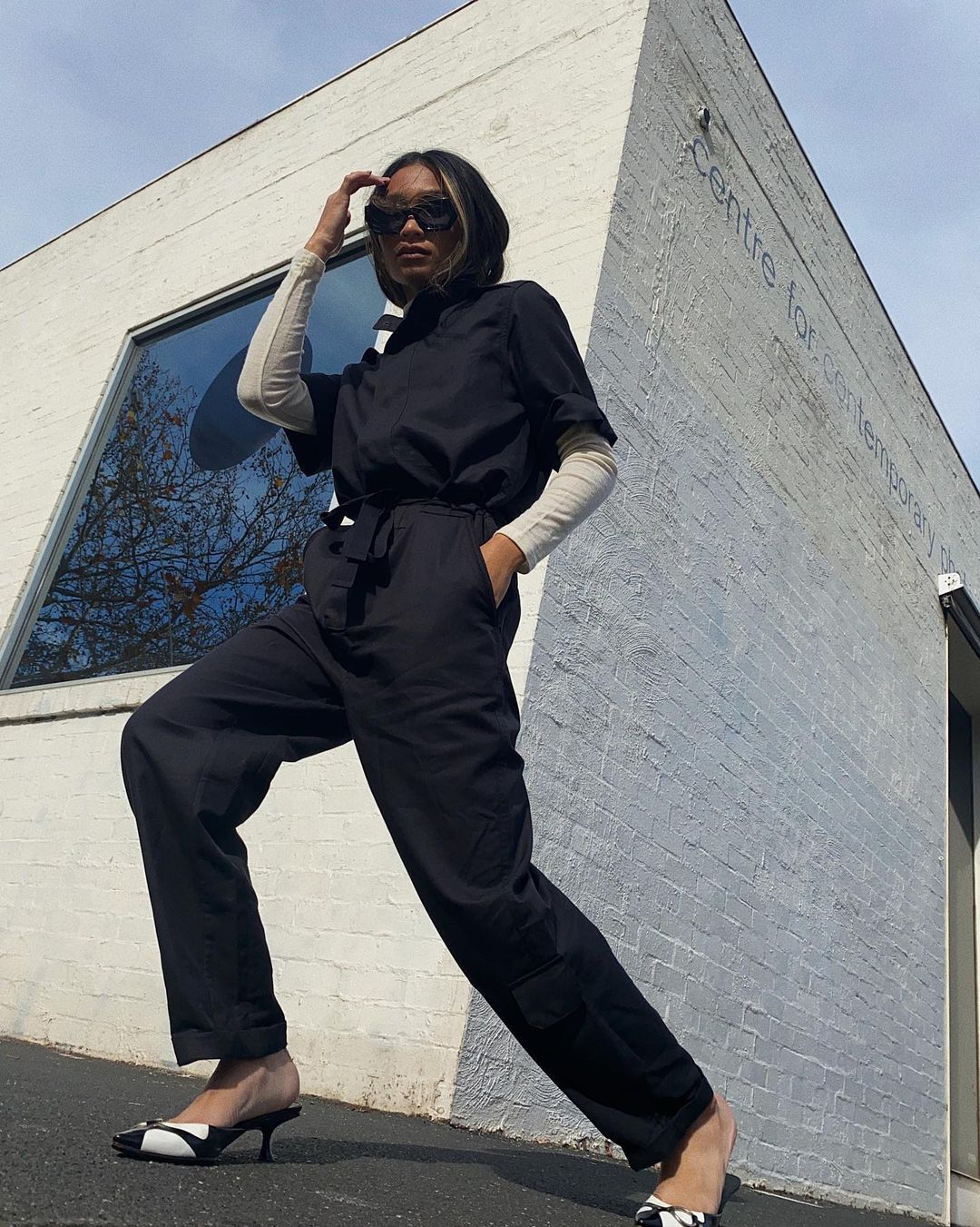 Stylish adult woman in black jumpsuit and patterned mules strikes a pose outdoors against a modern architectural backdrop.