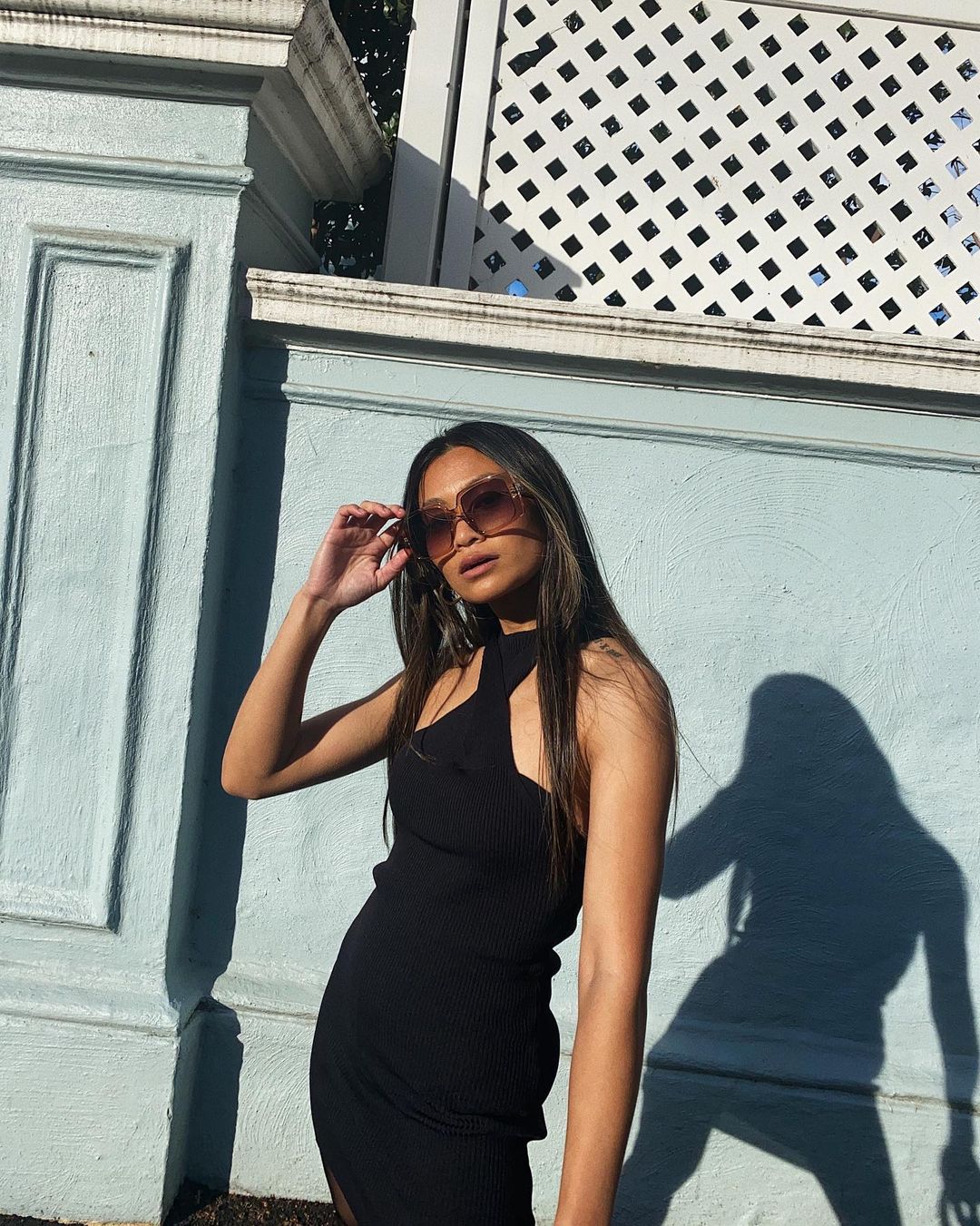 Attractive young woman in a stylish black dress and sunglasses poses in an urban setting with a light blue wall background.