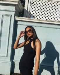 Woman in black halter dress wearing oversized sunglasses outdoors against a light blue textured wall.
