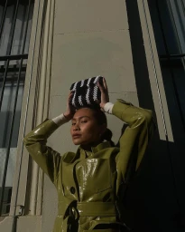 Woman in shiny olive green coat holding a black and white beaded bag above her head against an architectural building wall