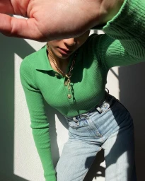 Woman in a vibrant green ribbed polo top and high-waisted denim jeans holding a hand up to block the camera.