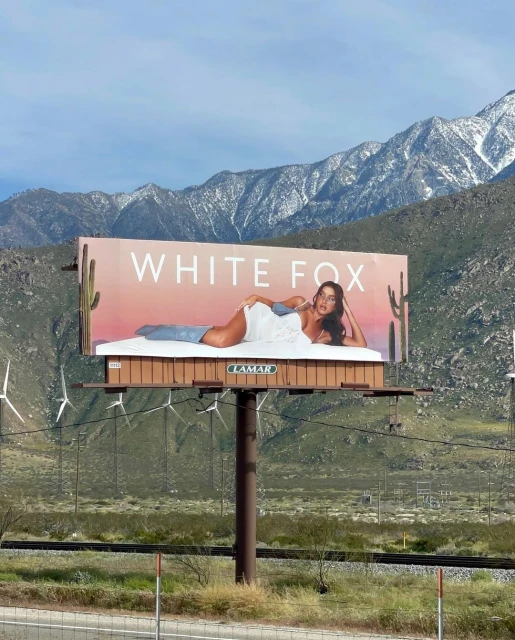 Billboard advertisement featuring a woman reclining on a bed, with the text 'WHITE FOX,' set against a background of mountains and windmills.