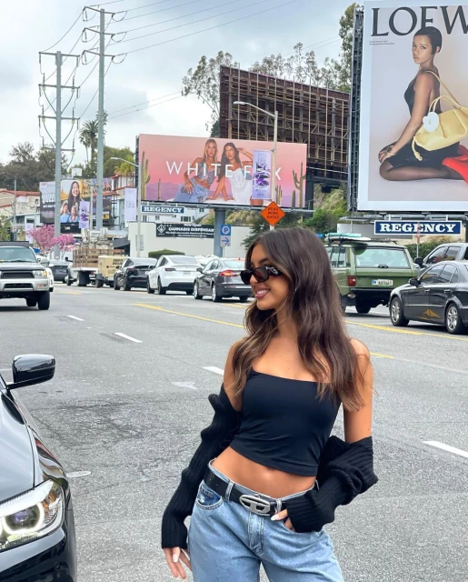 A fashionable woman in a black top and jeans stands on a city street with billboards, cars, and buildings in the background.