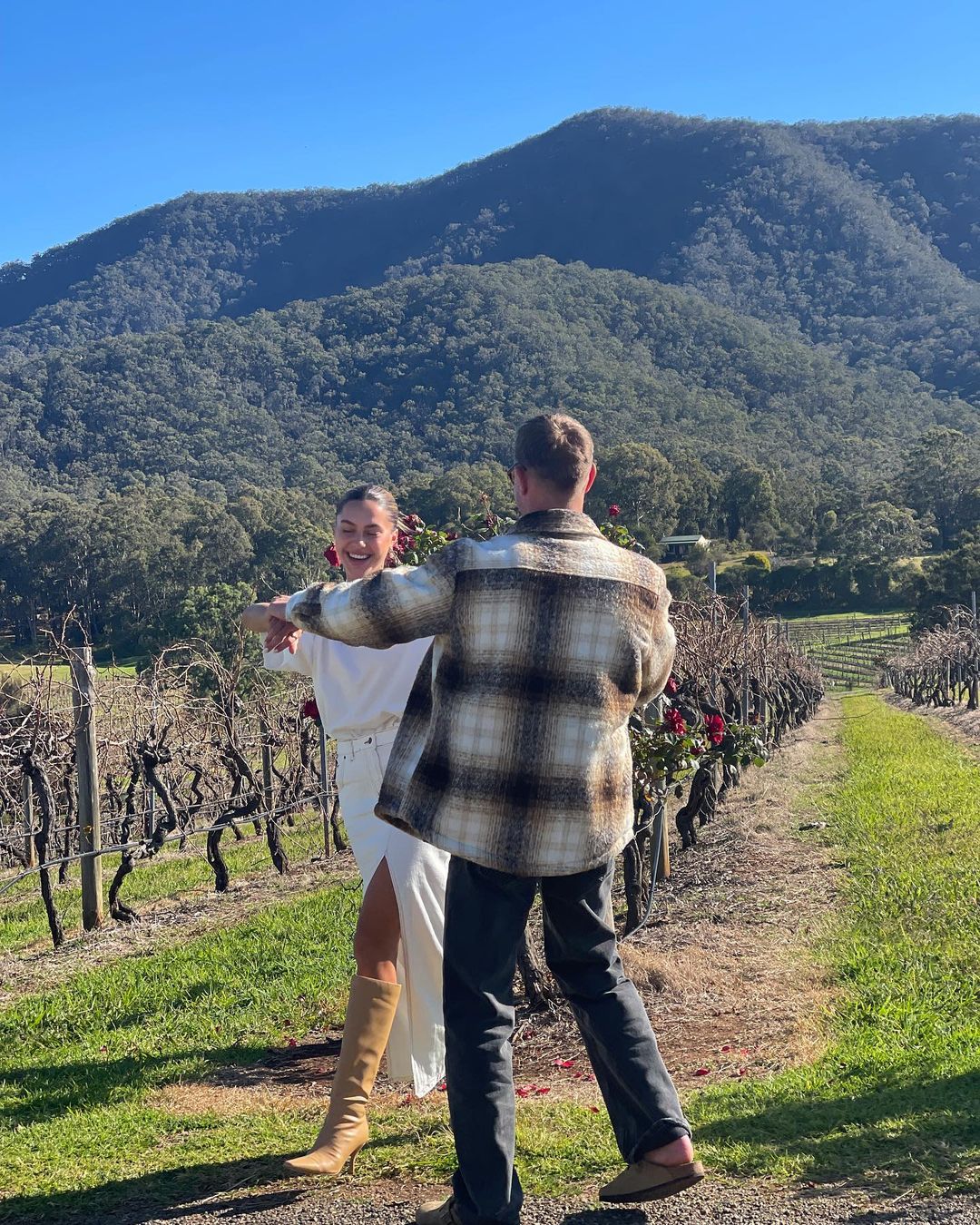 A couple stands in a vineyard with mountains in the distance. The woman wears a white dress and boots, while the man wears a flannel shirt.