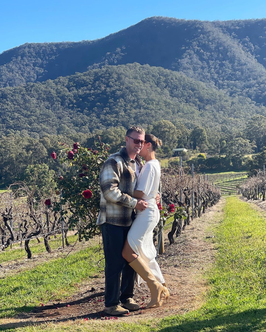 Couple embracing in a vineyard with a backdrop of tree-covered mountains under a clear sky on a sunny day, captured in natural light.