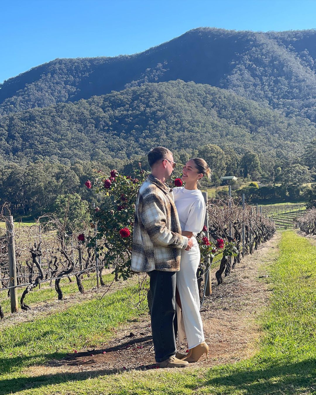 Couple poses in vineyard with mountains in the background. The woman wears white while the man is in a plaid jacket, under blue sky.