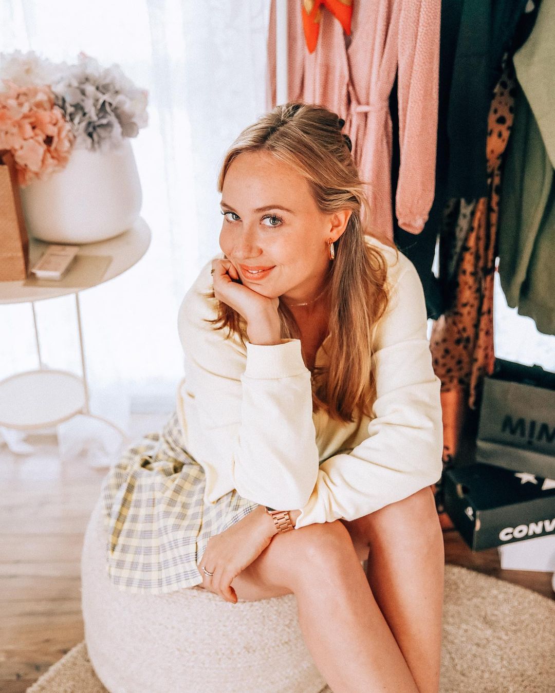 A young fair-skinned woman in casual yellow top and plaid shorts sits indoors on a white cushion. Accessories and clothing rack visible.
