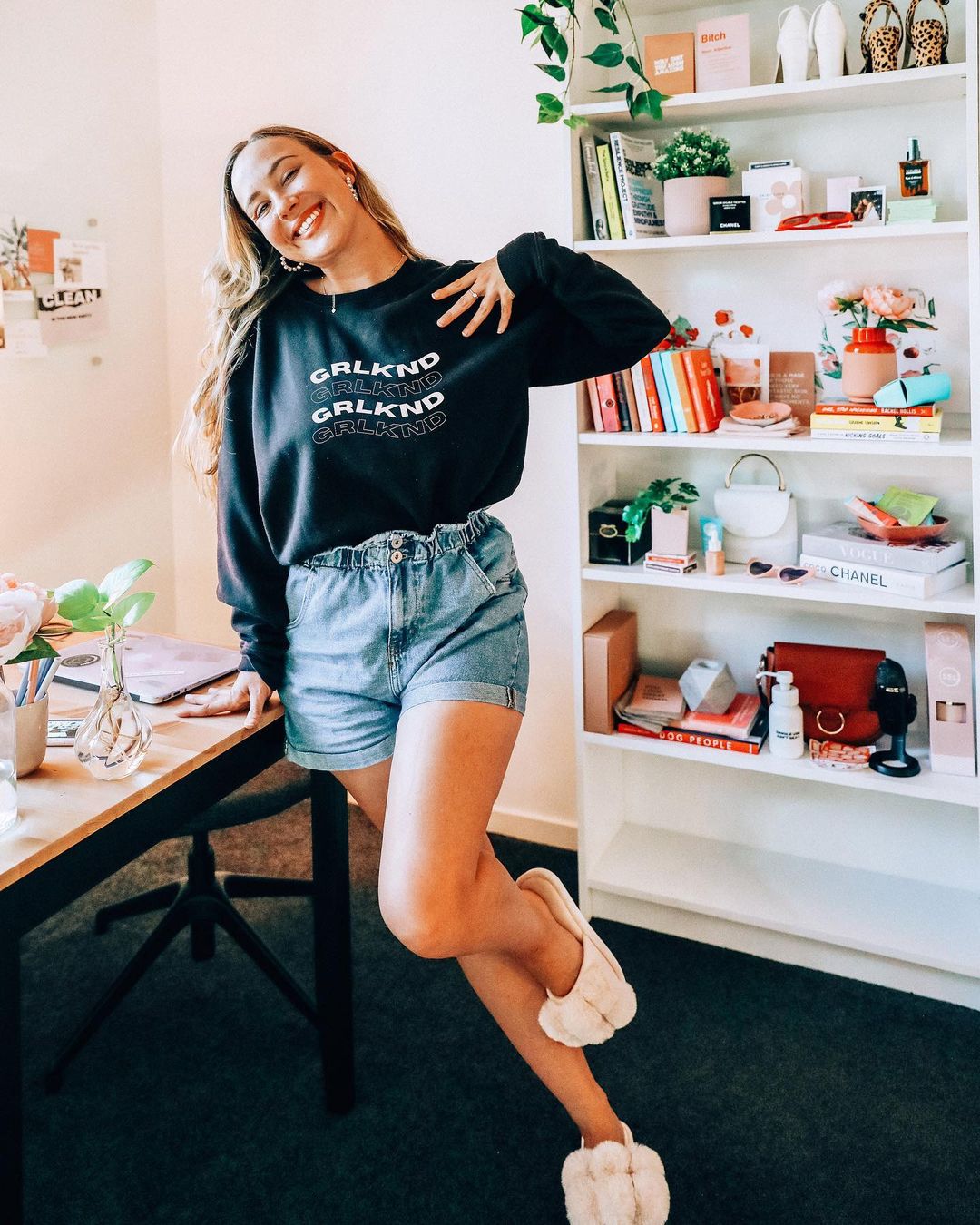 A young woman smiles while leaning against a desk with a laptop and flowers; a bookshelf filled with books and objects in the background.