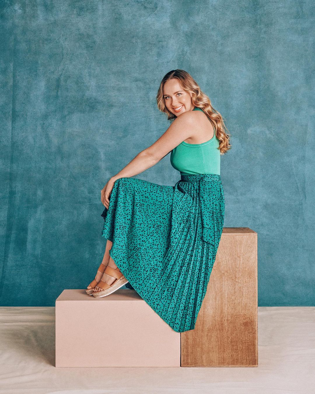 Blonde woman seated on blocks against a blue backdrop, wearing a green top and printed skirt, smiling and looking at the camera.