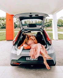 Woman in pink dress and straw hat sitting in the open trunk of a silver hatchback car