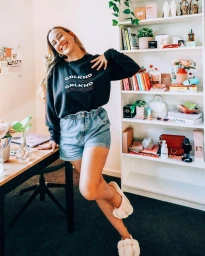 Content creator Milly Bannister posing by a desk in a home office wearing a black sweatshirt, denim shorts, and fluffy slippers.