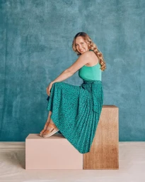 Smiling woman in green top and patterned pleated skirt seated on wooden blocks against a textured teal backdrop