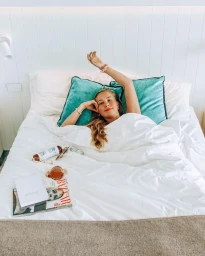 A woman lying in a white bed with teal pillows, raising her arm with a magazine and cosmetics resting on the sheets.