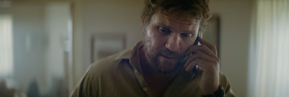 Man indoors talking on a cell phone wearing a casual khaki shirt, fair skin, and an athletic watch. Looking at camera.