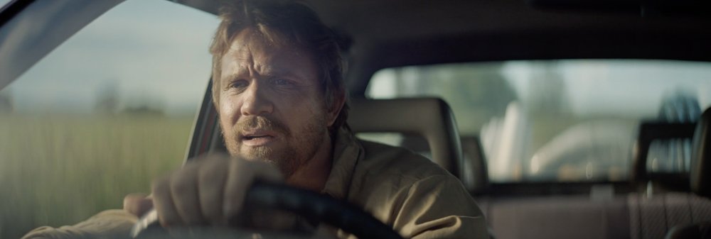 Adult man driving an older car on a rural road. The man is wearing a light-colored shirt and appears concerned in the driver's seat.