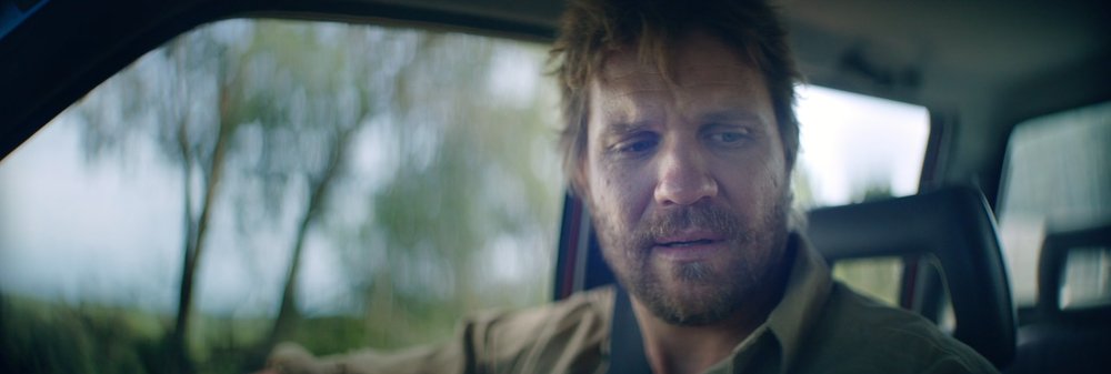 Close-up of a bearded man driving a car with trees blurred in the background, focusing on the driver's expression and vehicle interior.
