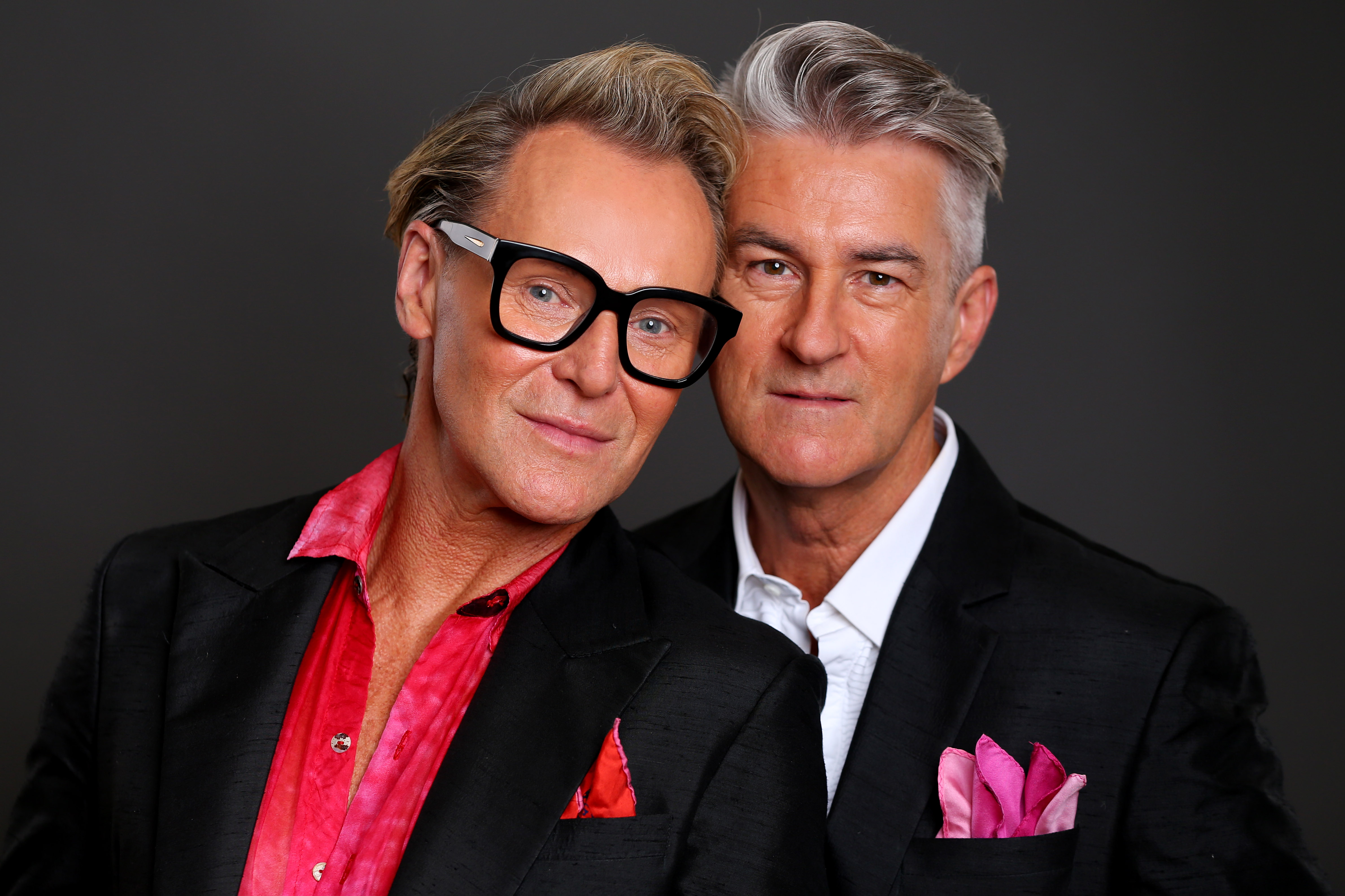 Portrait of two adult men, one with blonde hair and glasses, and one with grey hair, wearing black suits and smiling in a studio setting.