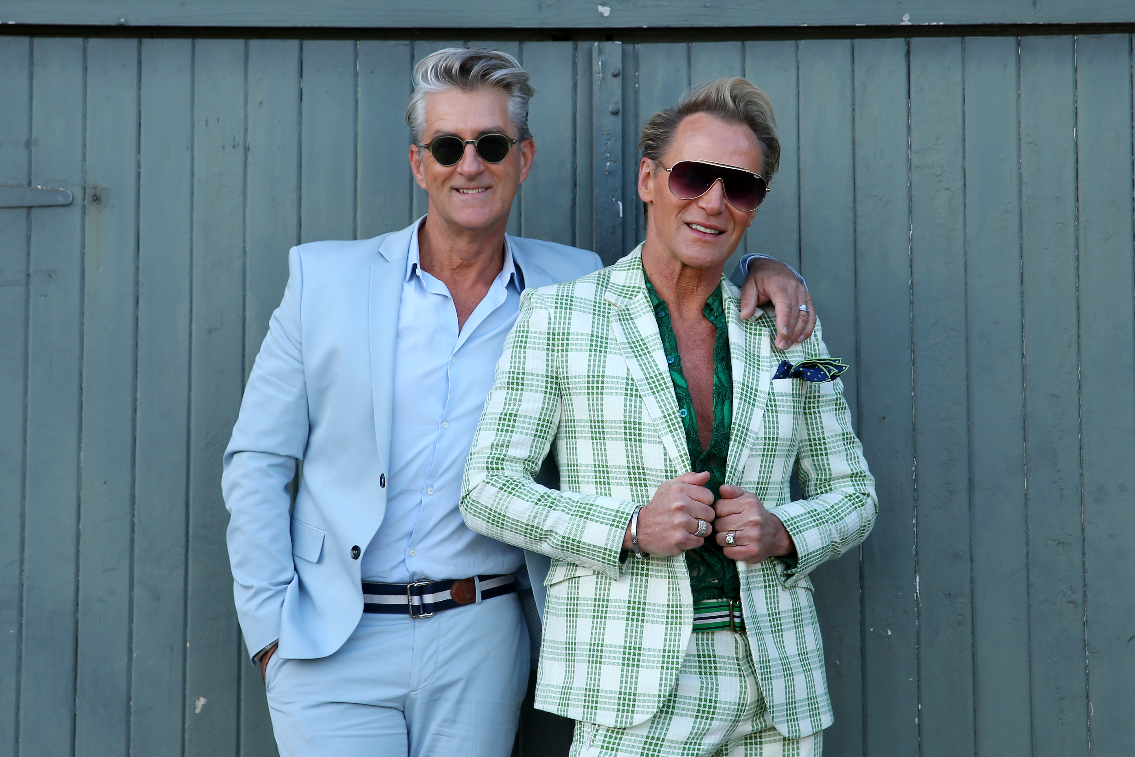 Two adult males pose outdoors wearing stylish suits and sunglasses in a medium shot against a grey wall for fashion or lifestyle content.