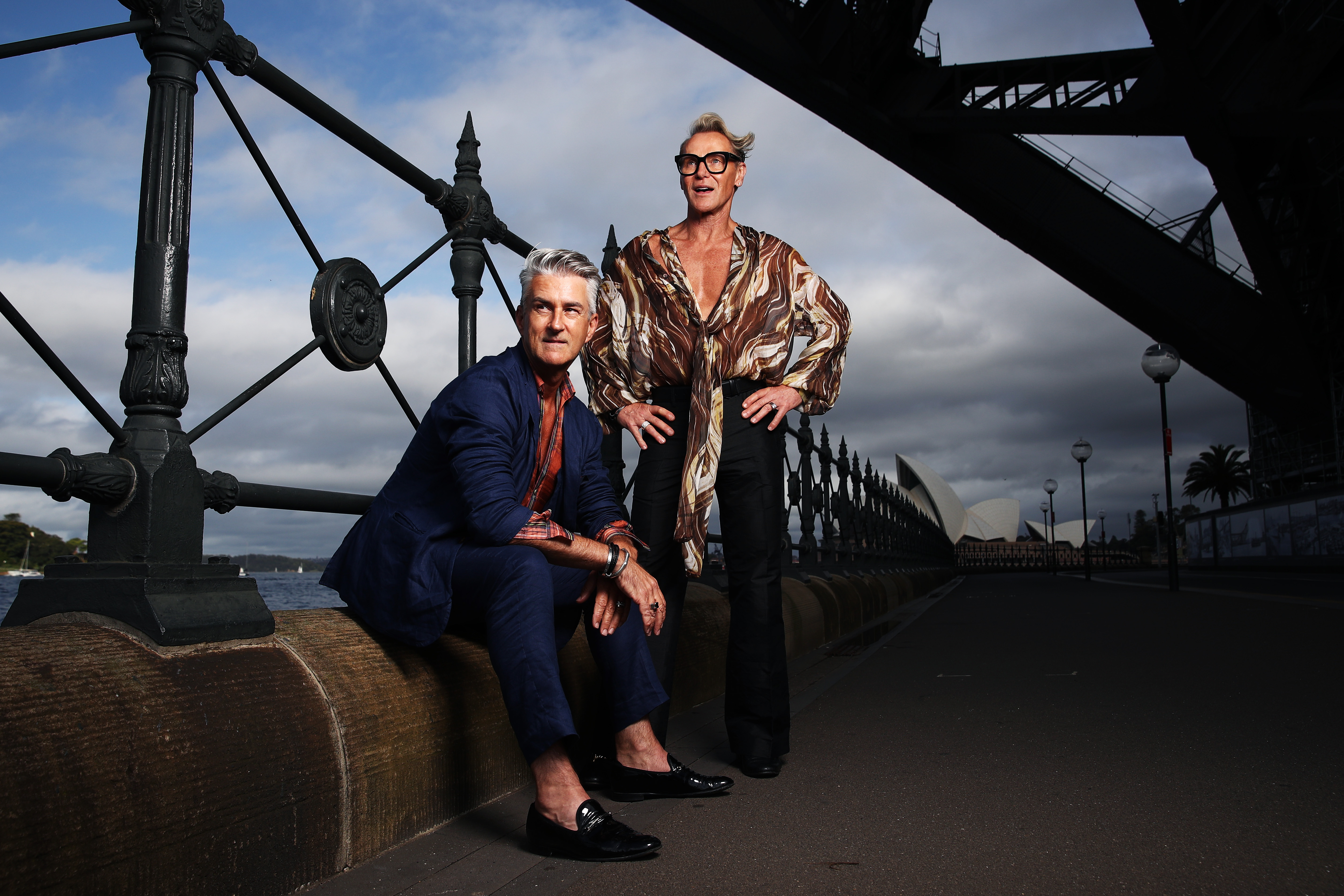 Two stylish adult males pose outdoors near a bridge, one seated and wearing a blue suit, the other standing in a patterned shirt.