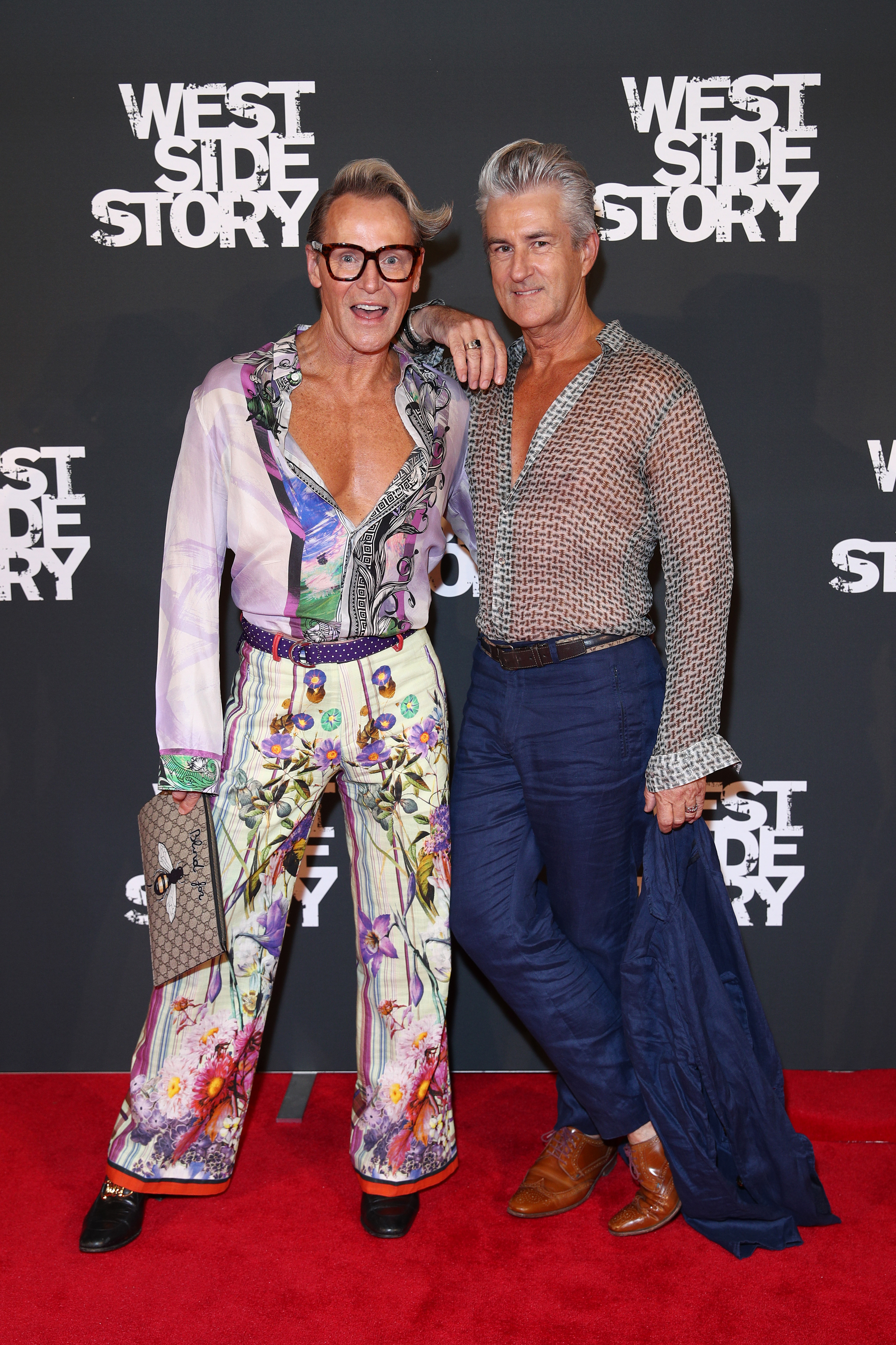 Two men pose at the West Side Story premiere, one with a patterned top and pants, the other in sheer shirt and navy trousers.