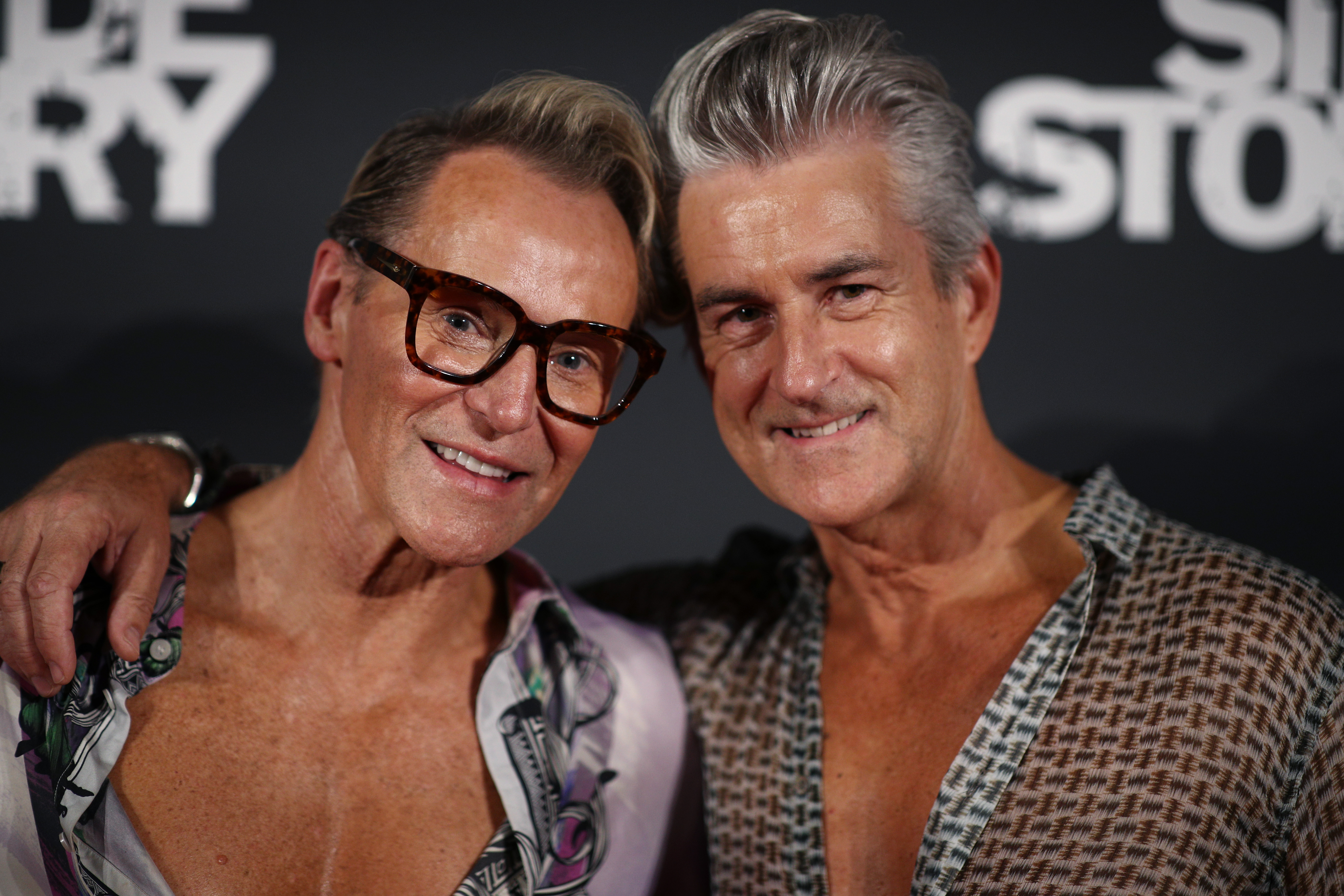 Two middle-aged adult males smiling and posing closely together at an indoor event in front of a backdrop with text. Focus on the pair.