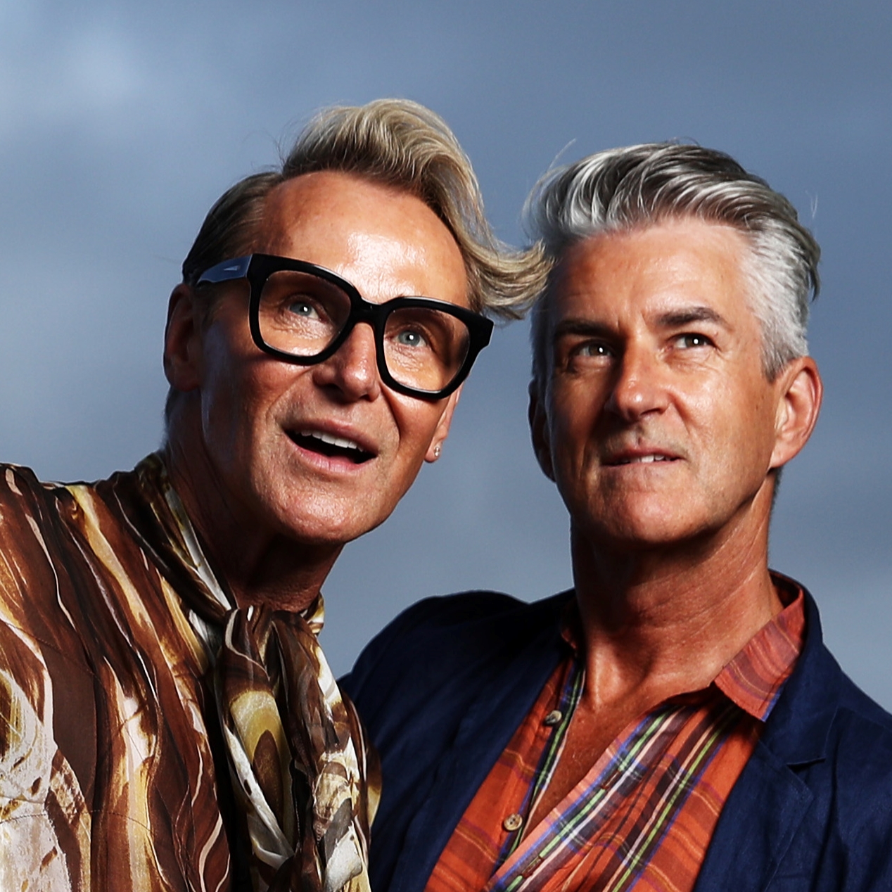 Close-up of two adult men in stylish shirts. One wears glasses, looking up and smiling; the other, in a blazer, looks off to the side.