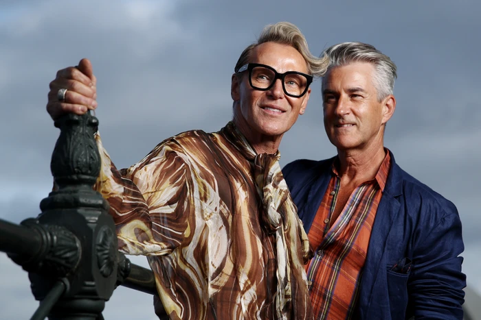 Two middle-aged men standing outdoors outdoors in Sydney, one wearing black glasses and a patterned silk shirt leaning against a railing.