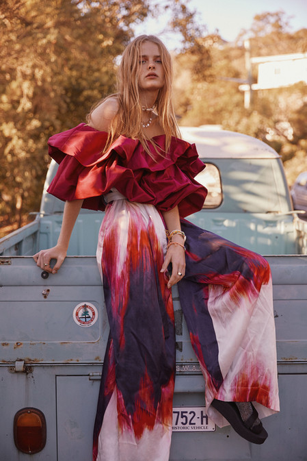 A blonde female fashion model poses outdoors sitting on the back of a vintage truck, wearing a red ruffled top and patterned pants.