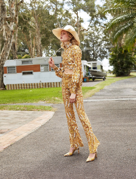 A woman in a gold patterned outfit and a straw hat poses on a road with trees, trailers, and green grass in the background.