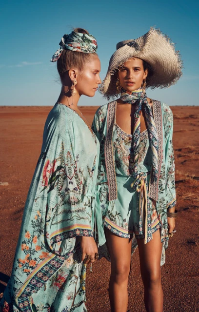 Two women in fashionable, floral-patterned outfits, one in a sunhat, pose in a desert landscape under a clear sky. Fashion editorial.
