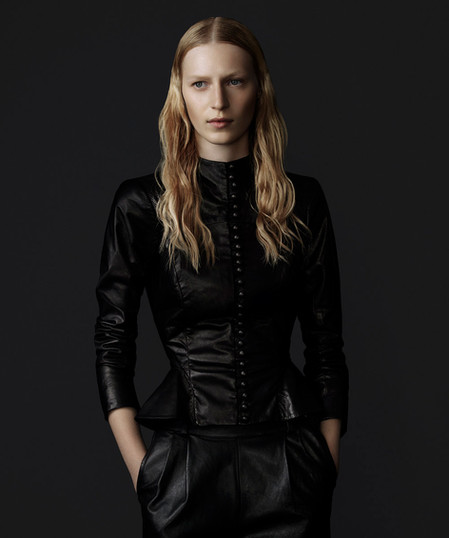 Elegant woman in a dark photo studio wearing a black leather peplum jacket with decorative buttons and black slacks, posing with hands in pockets.