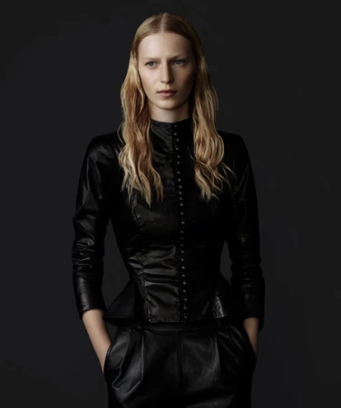 Elegant woman in a dark photo studio wearing a black leather peplum jacket with decorative buttons and black slacks, posing with hands in pockets.