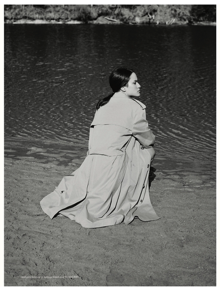 Model wearing a long trench coat crouching on a sandy beach near water styled by Nadene Duncan