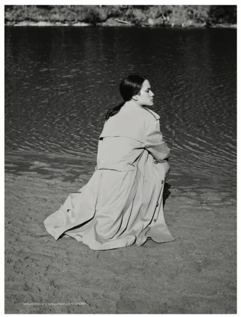 Model wearing a long trench coat crouching on a sandy beach near water styled by Nadene Duncan