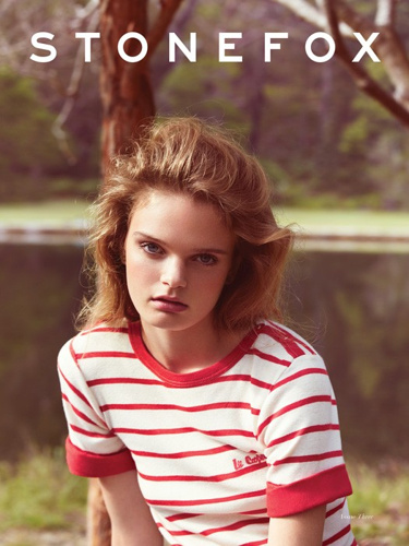 Young woman gazes at the camera, wearing a striped t-shirt in an outdoor setting with trees, creating a natural, fashion-focused scene.