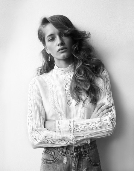 Portrait of a woman wearing a white lace high-neck blouse and denim jeans against a plain background