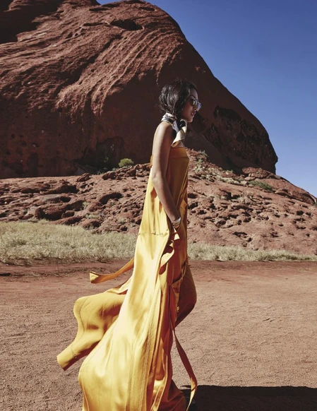 Woman in flowing gold dress walks across a red dirt landscape with a large red rock formation under a clear blue sky.