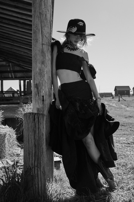 A woman in a black skirt and top, wearing a hat and boots, is leaning against a wooden post in a rural setting in this black-and-white photo.