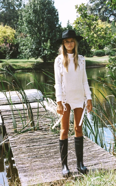 A blonde woman with a hat stands on a wooden dock near a pond, wearing a long, white knit sweater dress and black rain boots in nature.