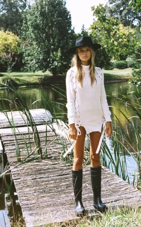A blonde woman with a hat stands on a wooden dock near a pond, wearing a long, white knit sweater dress and black rain boots in nature.