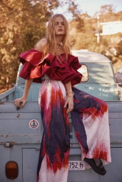 Model in off-the-shoulder red top and tie-dye trousers leaning against a vintage pickup truck outdoors.