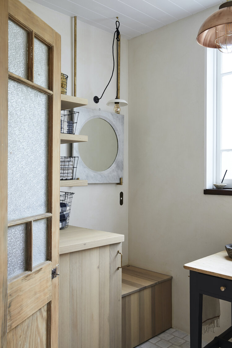 Bright, minimalist bathroom featuring an open wooden door, mirror, shelves, cabinets, hanging pendant light, and a neutral color scheme.