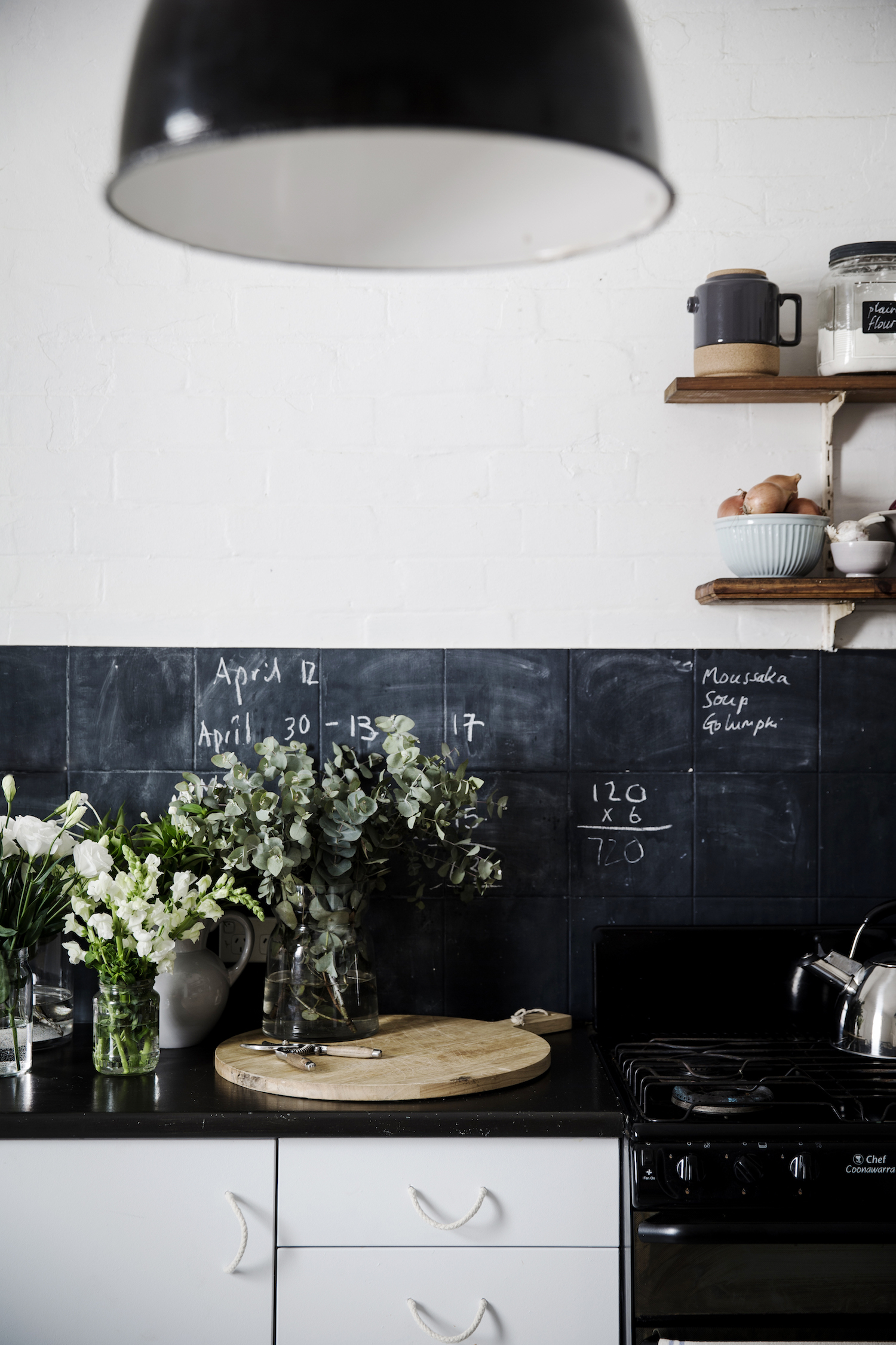 Kitchen interior featuring white cabinets, chalkboard tile backsplash with handwritten notes, flowers in vases, and kitchenware on shelves.