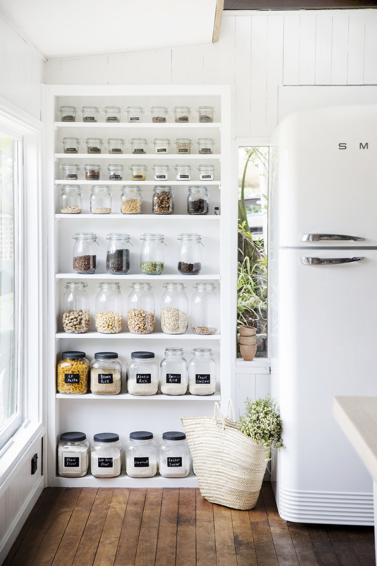 Pantry shelving filled with dry food goods inside clear jars, alongside a white SMEG refrigerator, in an indoor kitchen setting.