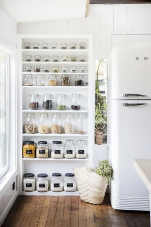 Pantry shelving filled with dry food goods inside clear jars, alongside a white SMEG refrigerator, in an indoor kitchen setting.