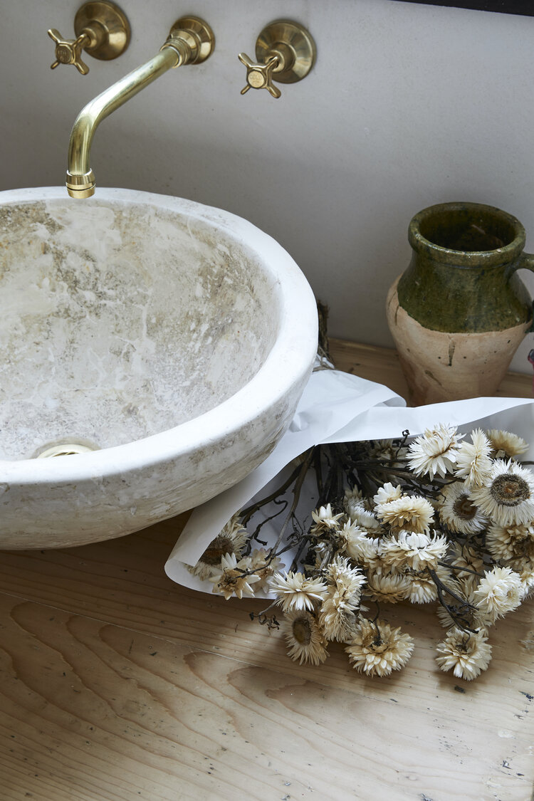 Close-up of a stone sink with brass fixtures, dried flowers, and a rustic vase on a wooden countertop in a well-lit bathroom.