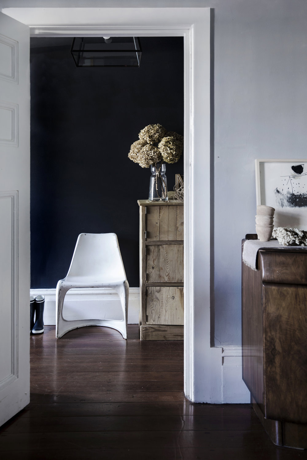 Interior view of a modern home featuring an open doorway, wooden floors, vintage dresser, white chair and dried hydrangeas in a vase.